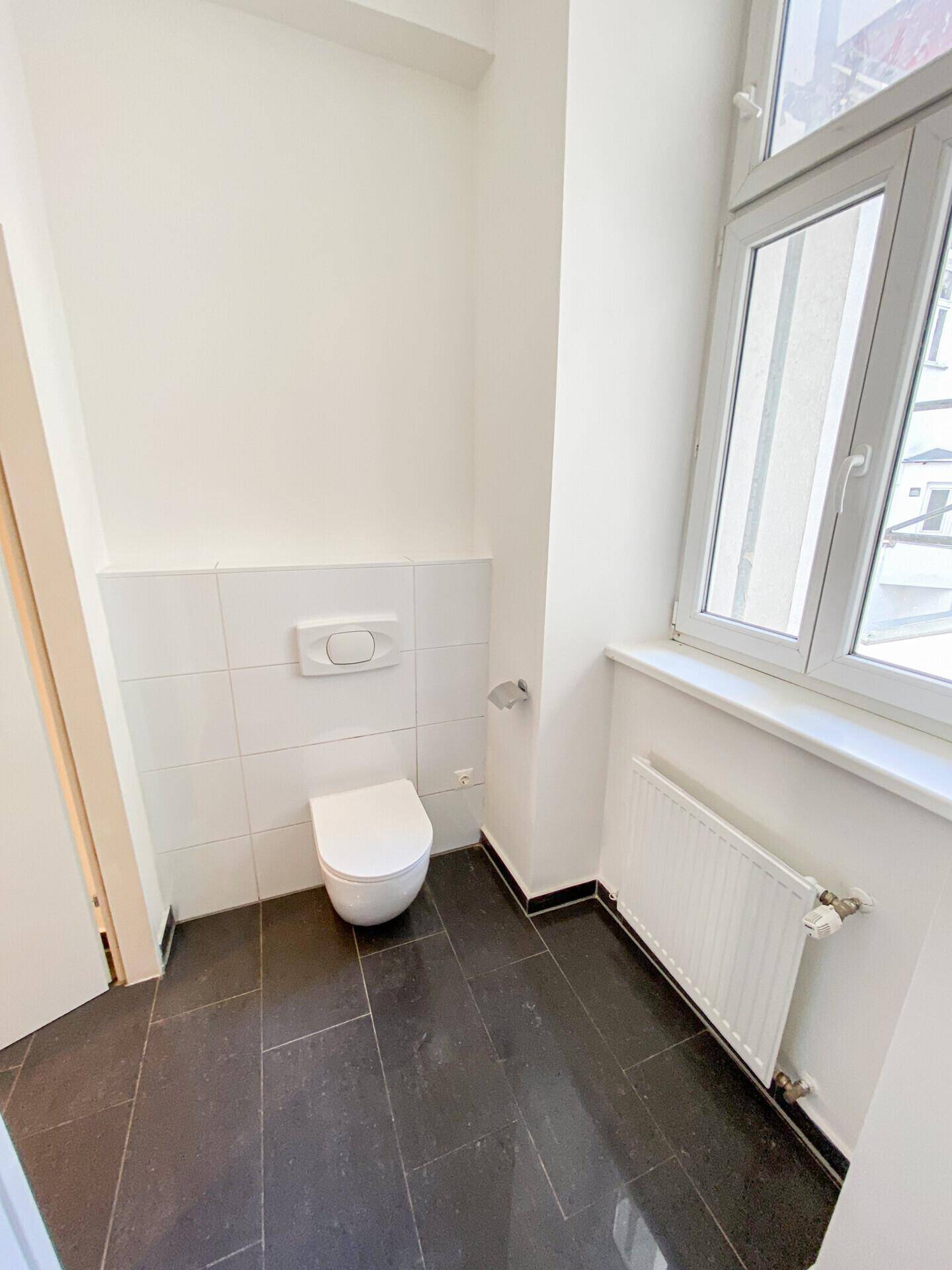 Separates WC mit wandhängender Toilette, Fenster und dunklem Fliesenboden.