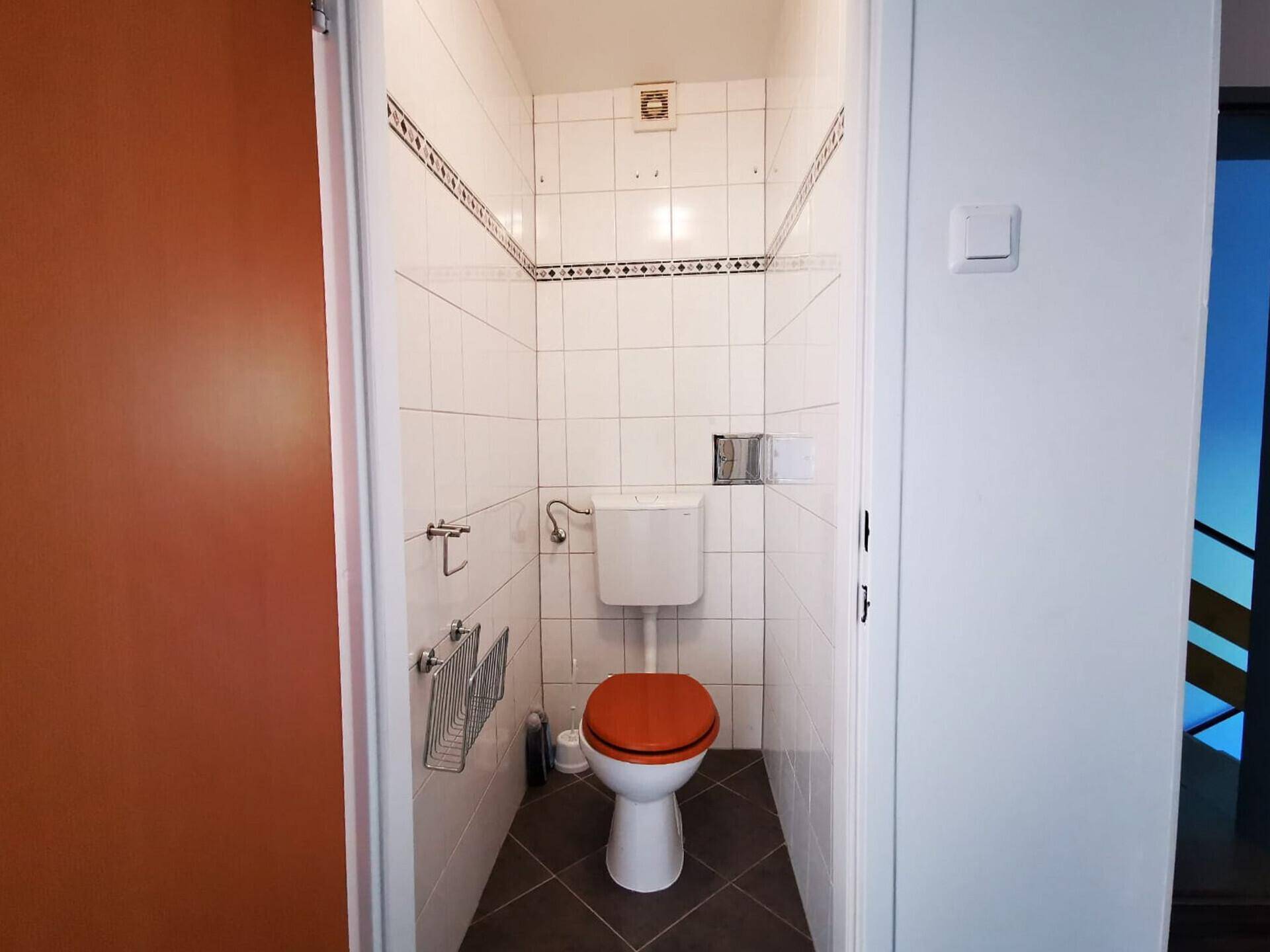 Kleines Gäste-WC mit weißen Fliesen, Toilette und orangefarbenem Toilettensitz.
