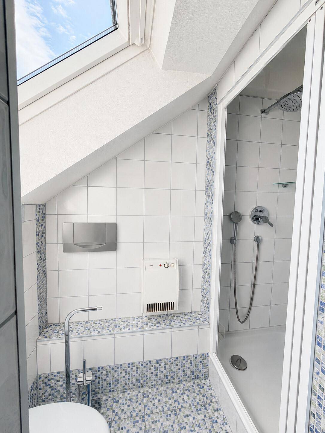 Duschbereich im Badezimmer mit Dachfenster, weißen Fliesen und blauen Mosaikdetails.