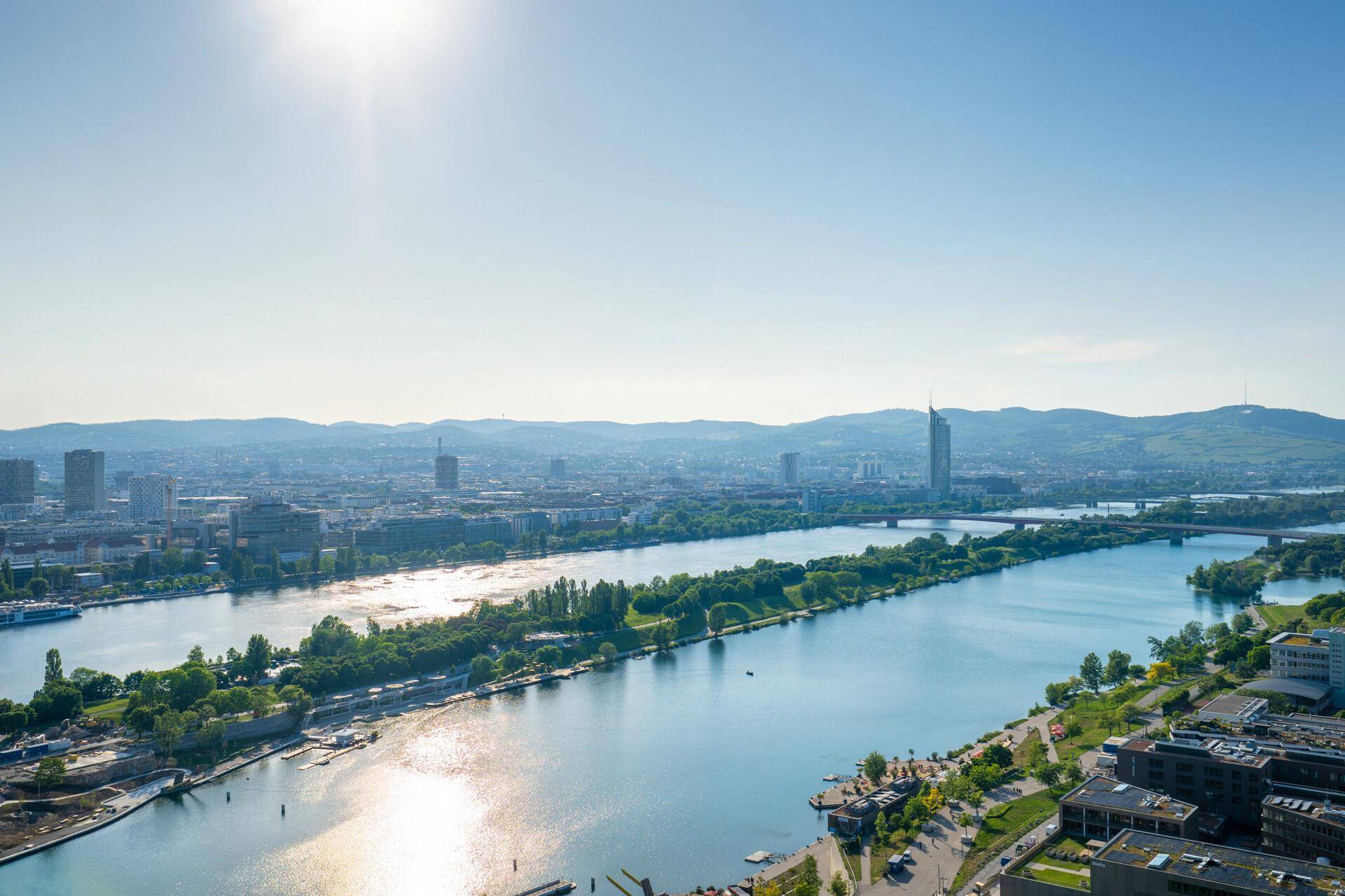 Panoramablick über eine weitläufige Stadtlandschaft mit Fluss, Brücken und Bergen am Horizont unter blauem Himmel.