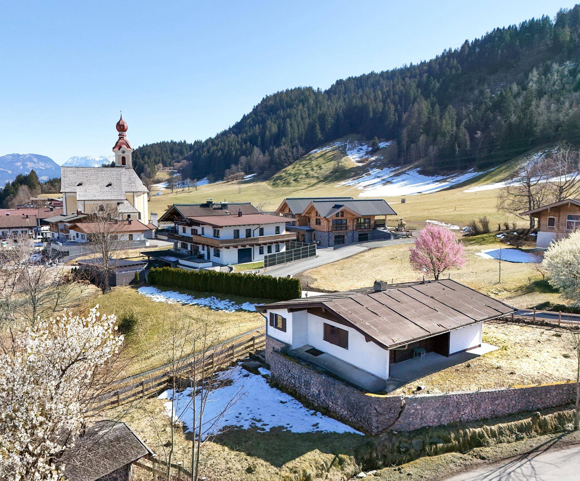 Panoramablick auf ein malerisches Dorf mit Kirche, umgeben von Hügeln und vereinzelten Schneefeldern.