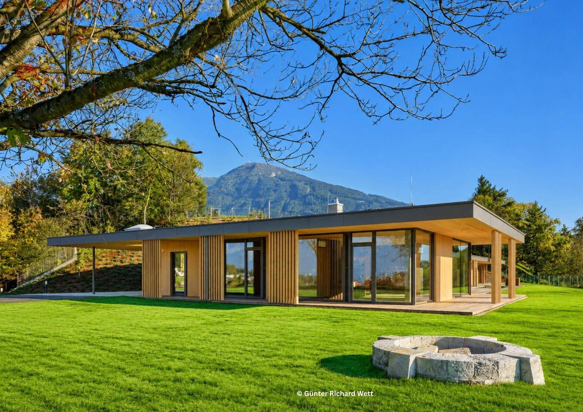 Moderne Villa mit Holzfassade, großem Garten und Feuerstelle vor malerischer Berglandschaft.