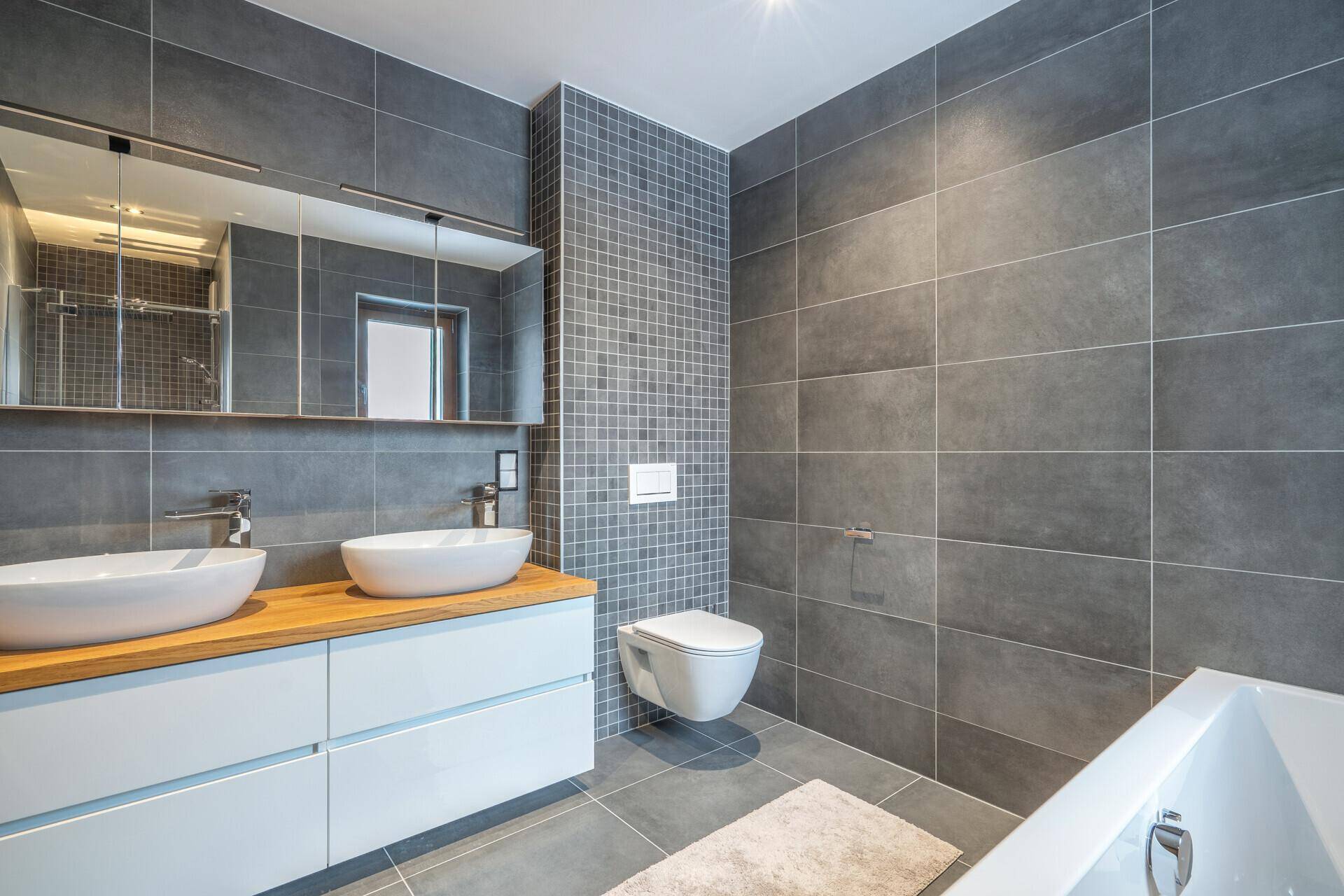 Modernes Badezimmer mit Doppelwaschbecken, grauen Fliesen, Badewanne und wandhängendem WC.