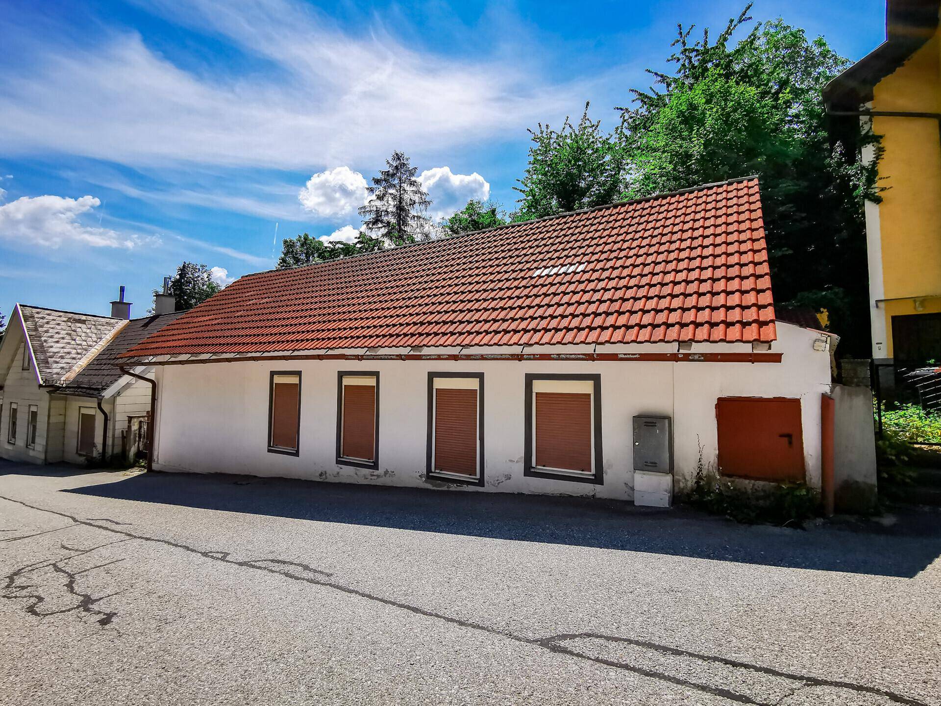 Lange weiße Hausfassade mit rotem Ziegeldach, mehreren Fenstern und geschlossenen Fensterläden.
