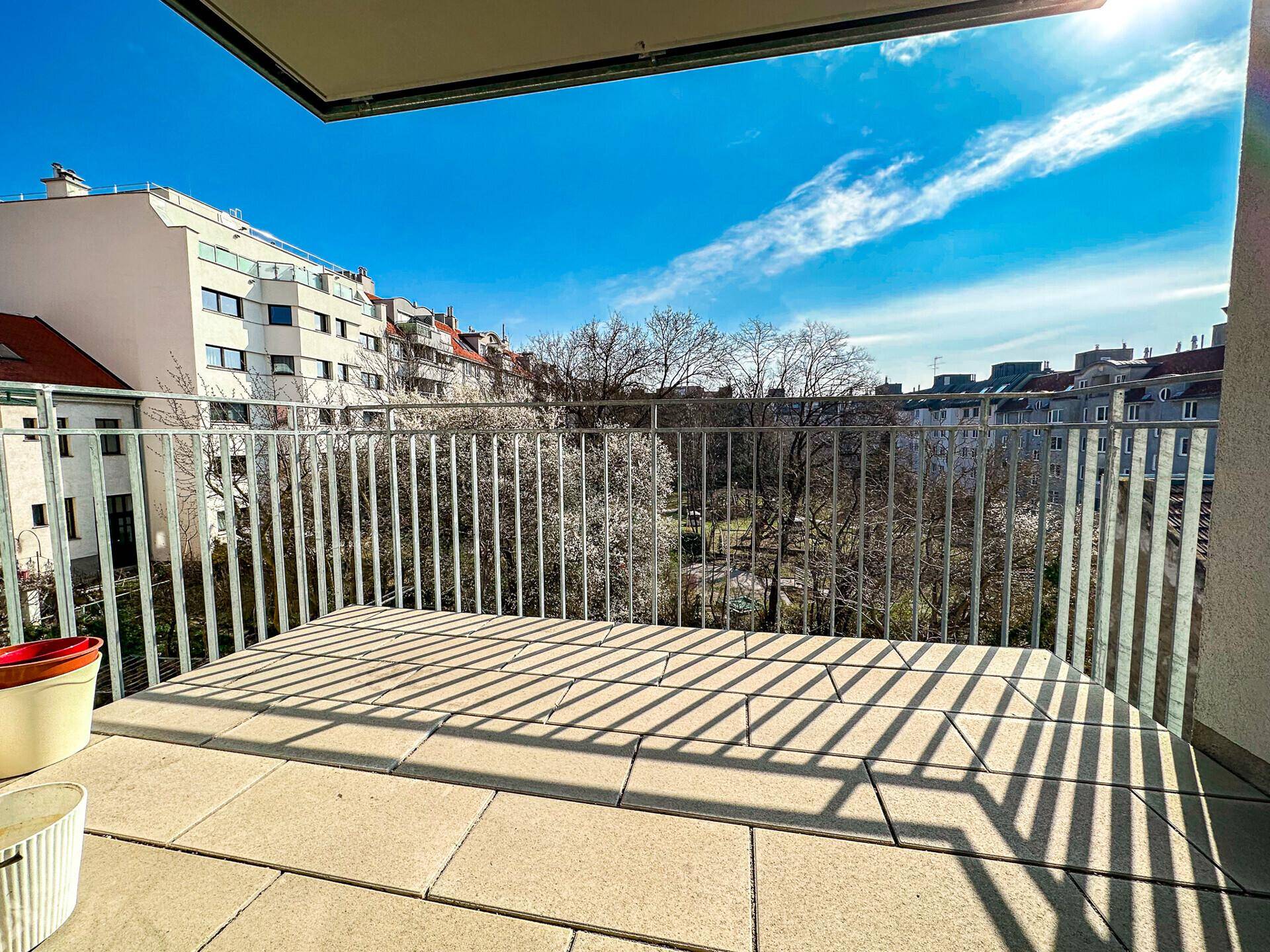 Sonniger Balkon mit Fliesenboden und Blick ins Grüne sowie auf umliegende Gebäude.