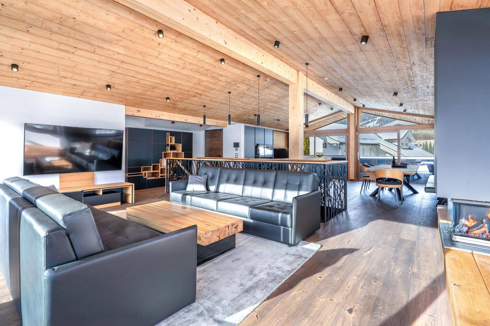 Geräumiges, offenes Wohnzimmer mit Holzbalkendecke, dunklen Sofas und Kamin in modernem Design.