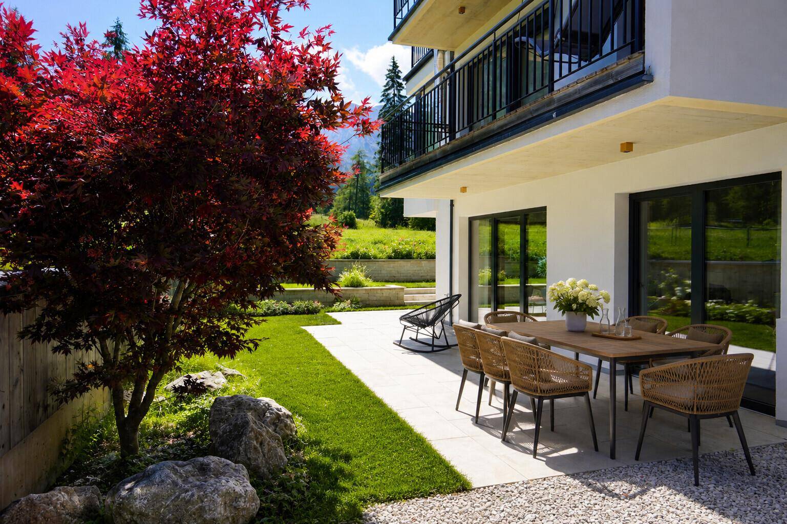 Sonniger Terrassenbereich mit Esstisch und Stühlen, umgeben von Grünflächen und einem roten Ahornbaum.