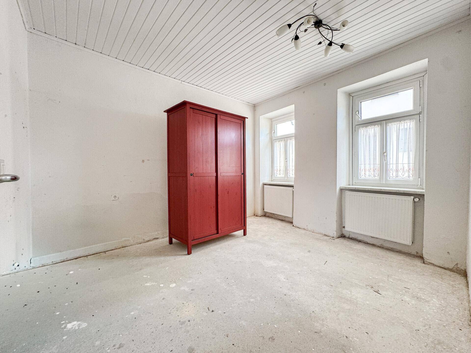 Helles Schlafzimmer mit Betonboden, zwei Fenstern und einem roten Kleiderschrank.