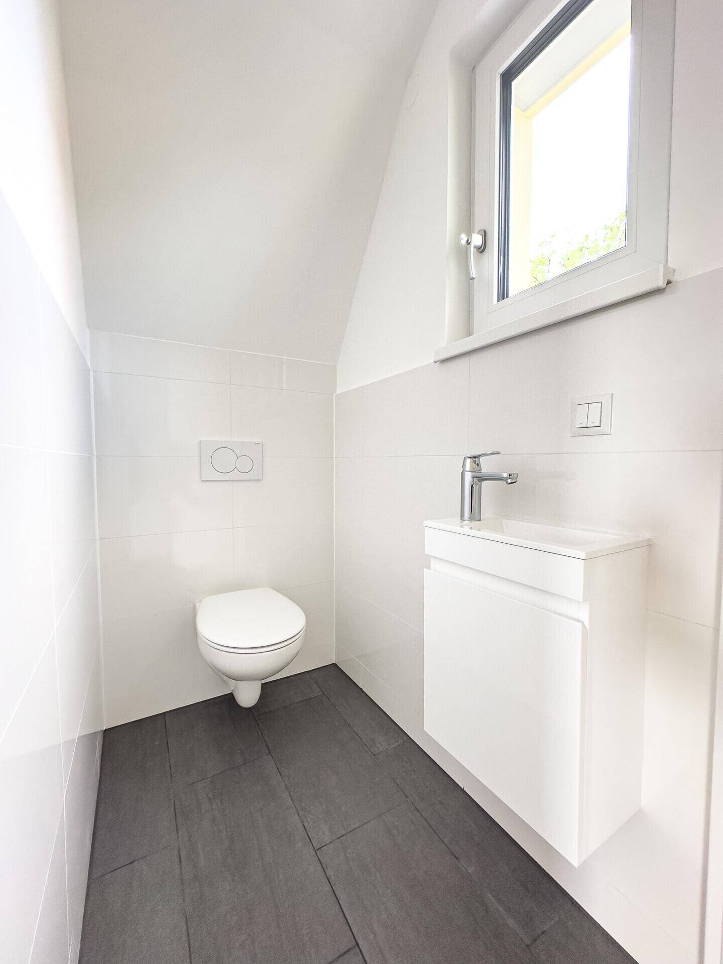 Helles Gäste-WC mit wandhängender Toilette, kleinem Waschbecken und Fenster für Tageslicht.