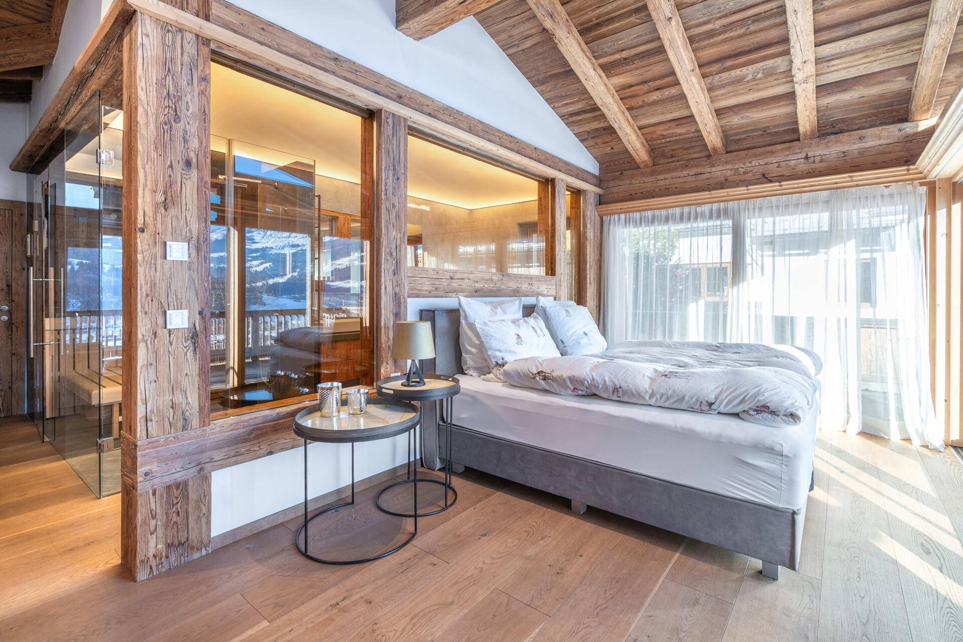 Gemütliches Schlafzimmer mit Holzbalkendecke, großen Fenstern und Zugang zu einem Wellnessbereich.