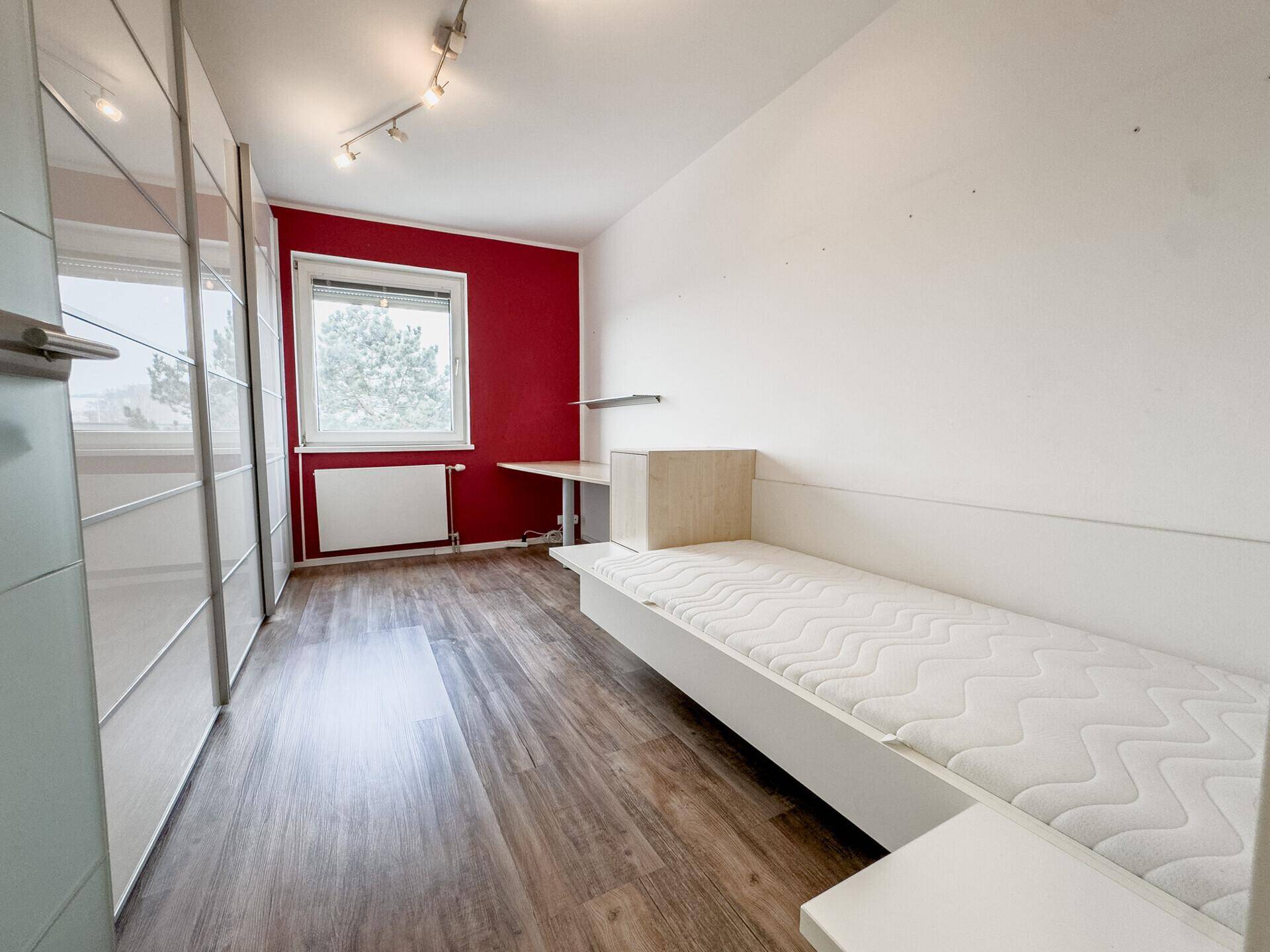 Helles Schlafzimmer mit Einzelbett, roter Akzentwand und großem Kleiderschrank.