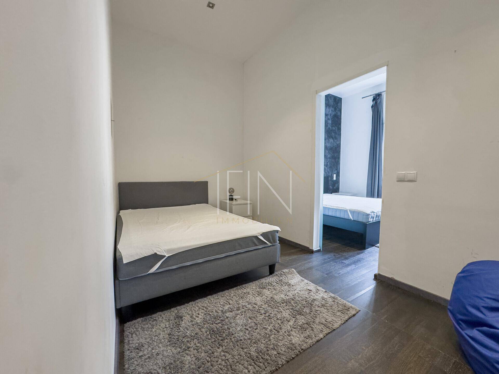 Kleineres Schlafzimmer mit Einzelbett und dunklem Holzboden, ein weiteres Zimmer ist im Hintergrund sichtbar.