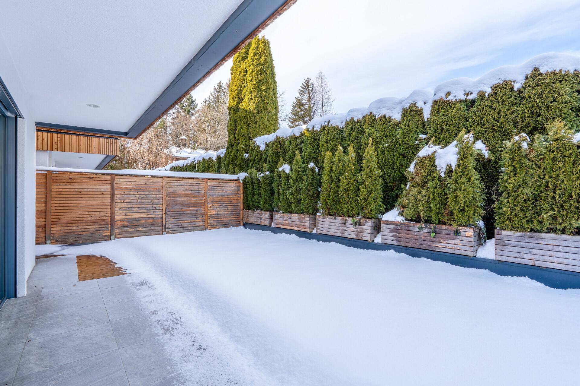 Überdachte Terrasse im Winter mit Schnee, grünen Hecken und Blick auf die Umgebung.