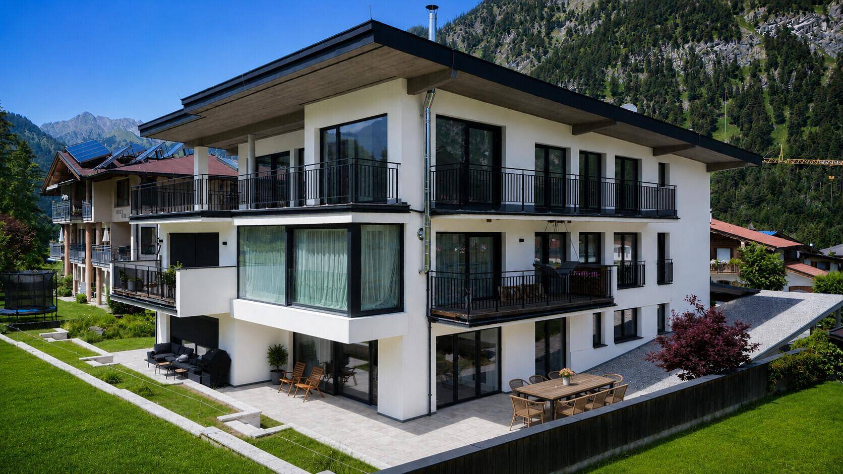 Elegantes modernes Gebäude mit großzügigen Glasflächen und Balkonen vor alpiner Bergkulisse.