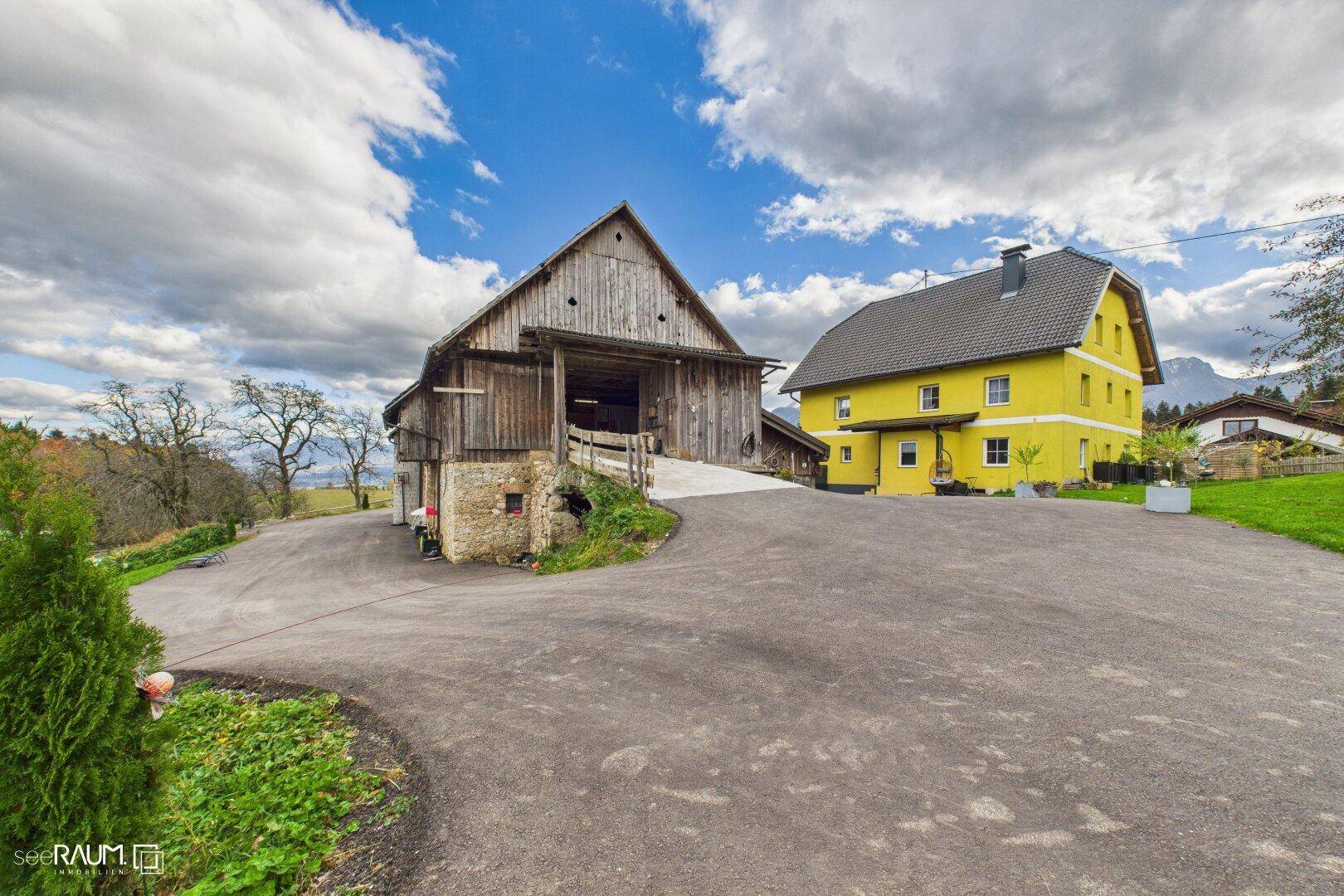 Weitläufiges Anwesen mit gelbem Wohnhaus und großem Holzstall vor Bergkulisse.