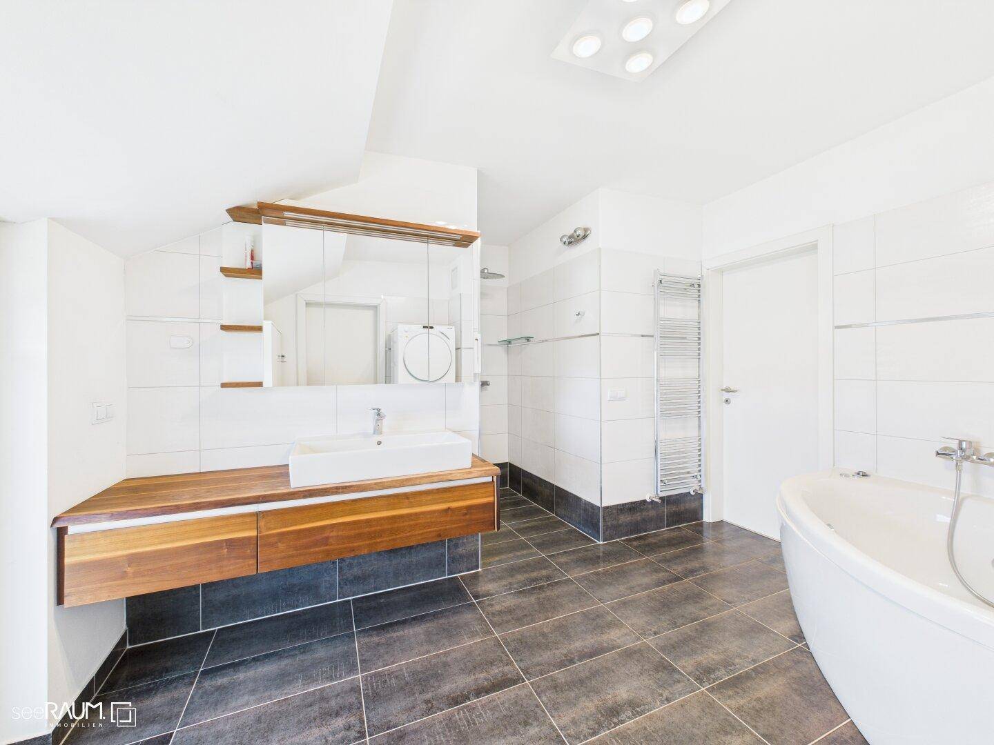 Modernes Badezimmer mit freistehender Badewanne, Holzwaschtisch und dunklen Fliesen.