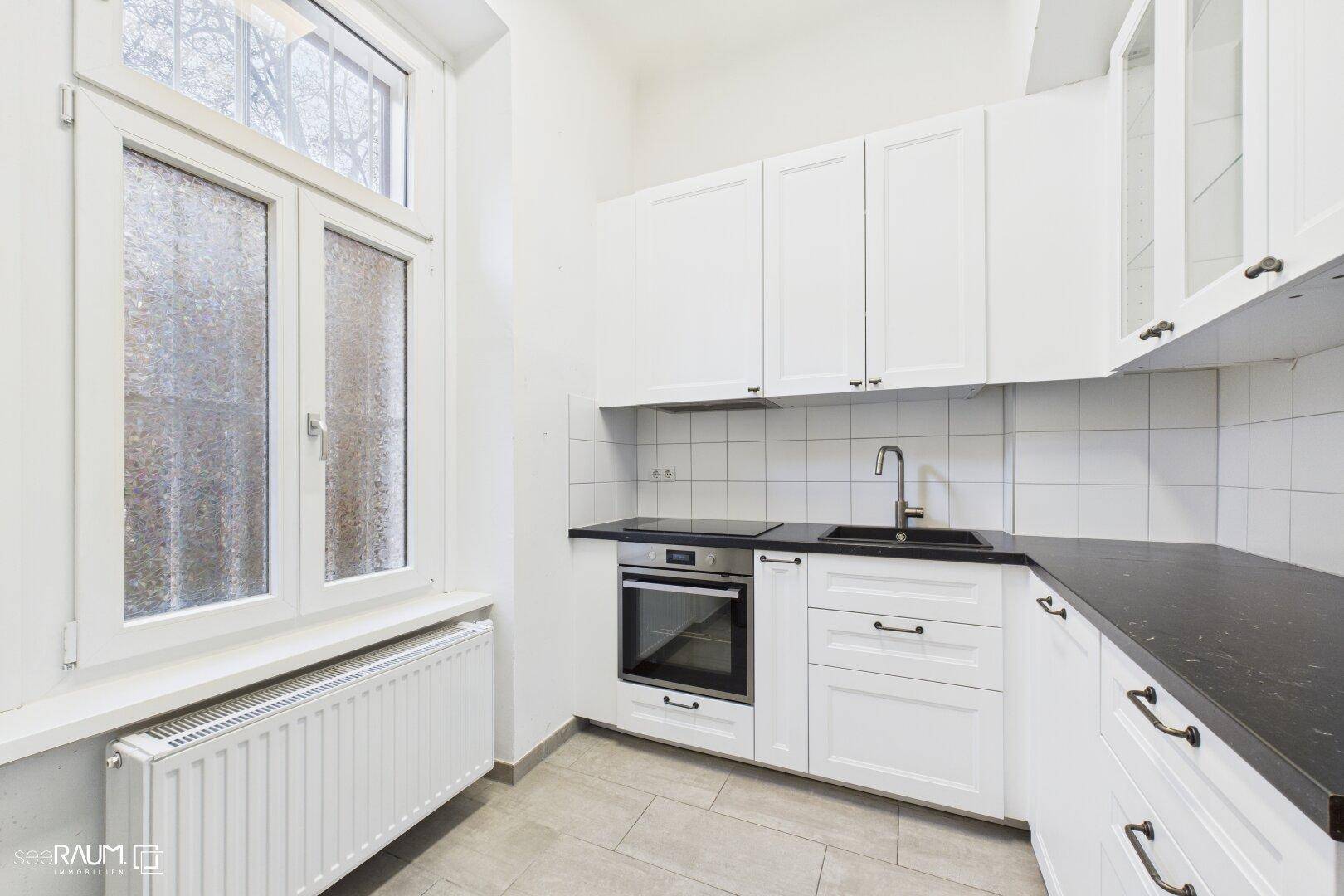 Moderne weiße Küche mit dunkler Arbeitsplatte, integriertem Backofen und Fenster.
