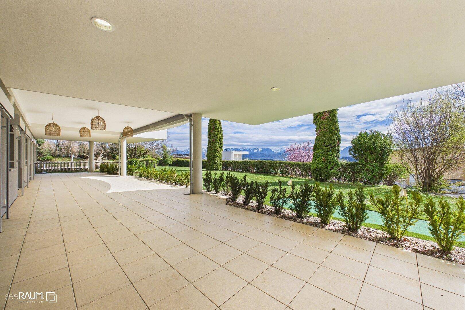 Überdachte Terrasse mit Fliesenboden und Zugang zu einem gepflegten Garten mit Blick auf die Landschaft.