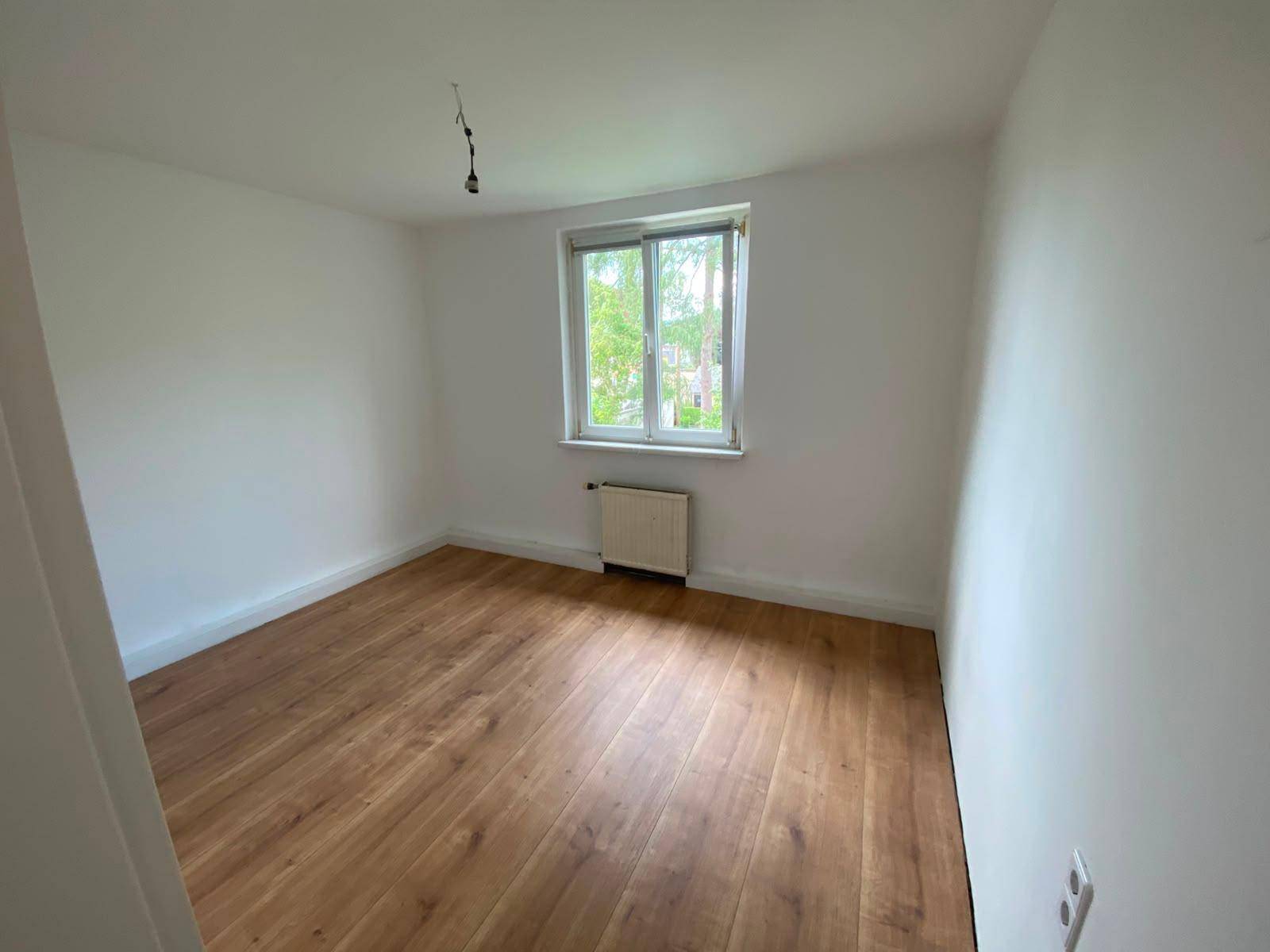 Leeres Zimmer mit Laminatboden, einem Fenster und weiß gestrichenen Wänden, bereit zur individuellen Gestaltung.