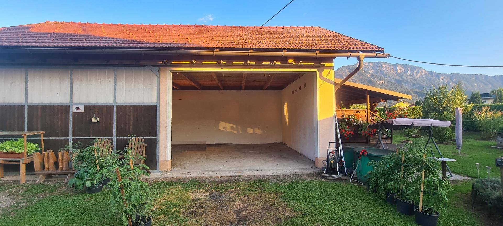 Blick in die offene Garage mit Betonboden, umgeben von einem Garten und einer Bergkulisse.