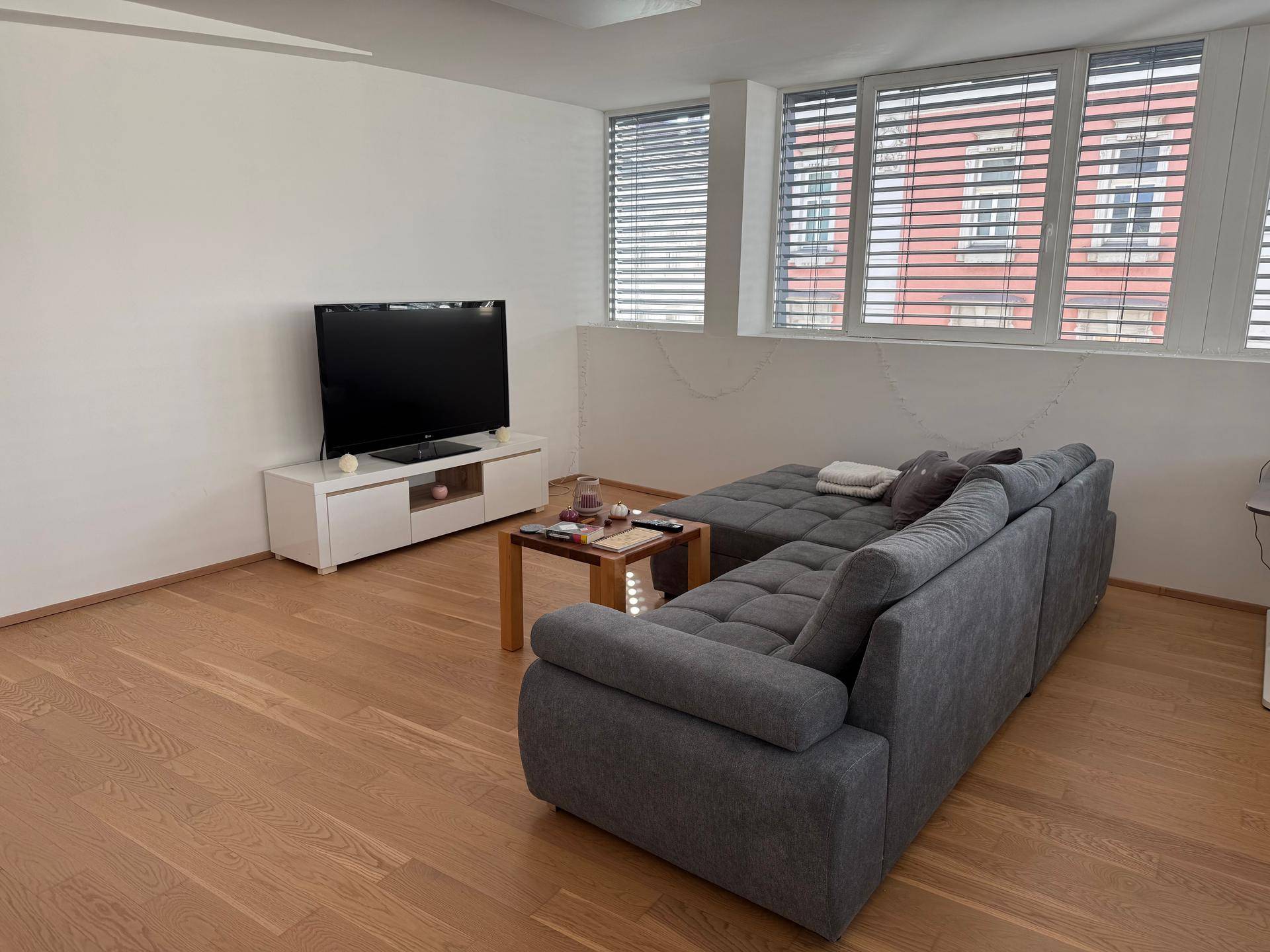 Gemütliches Wohnzimmer mit Sofa, Fernseher und Fenstern mit Jalousien für Privatsphäre.