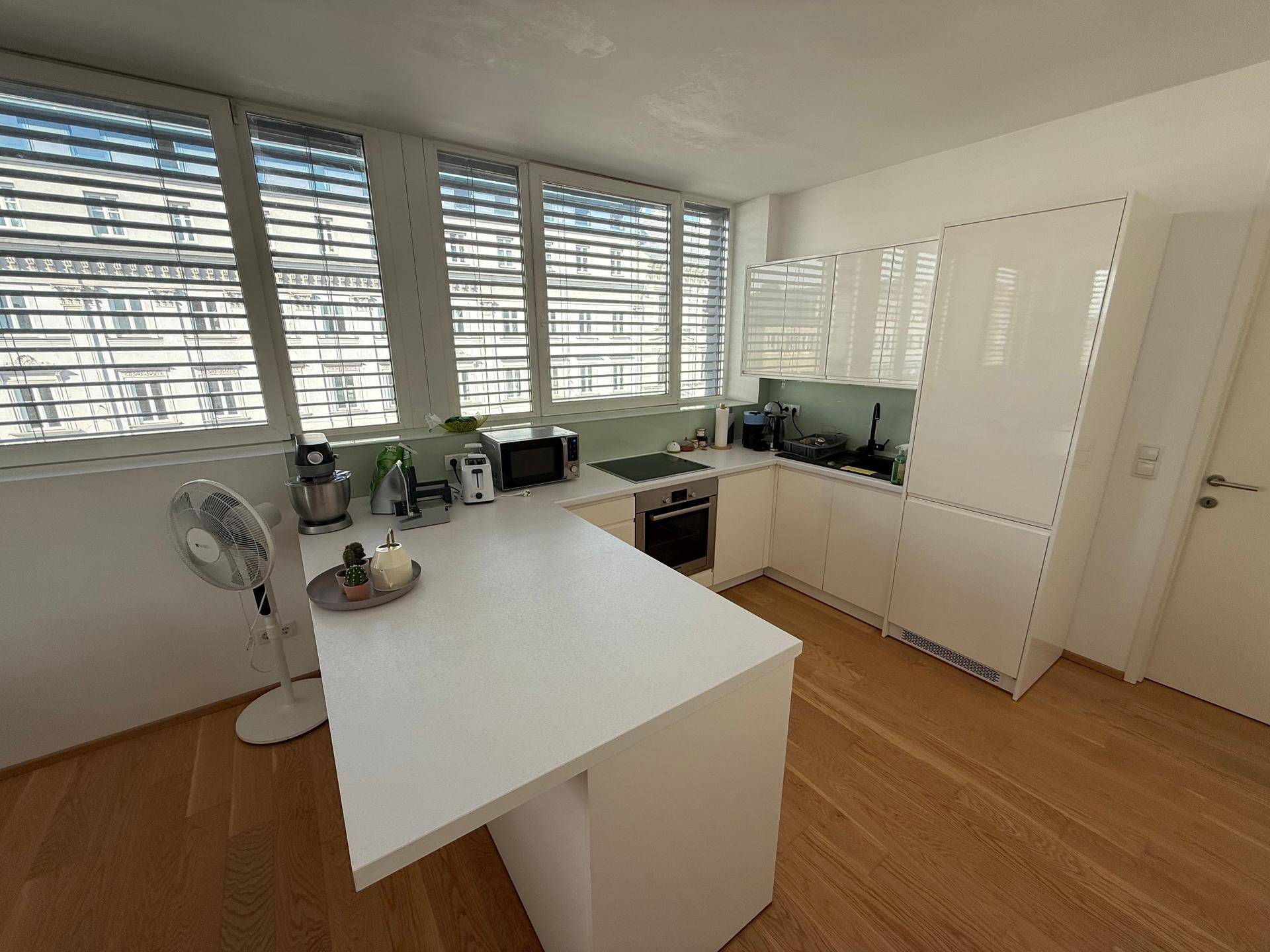Moderne weiße Küche mit Kücheninsel, integrierten Geräten und Fensterfront mit Jalousien.