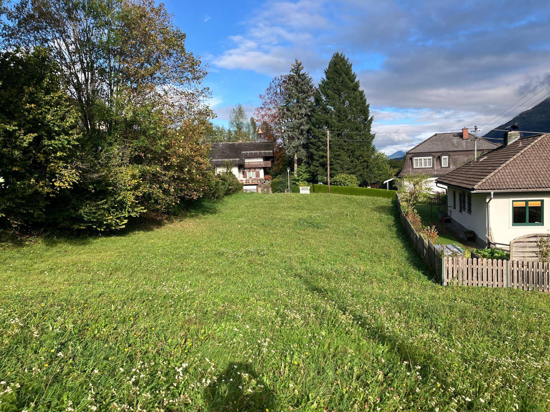 Weitläufiges grünes Grundstück mit einem Haus und Holzzaun auf der rechten Seite, umgeben von Bäumen.