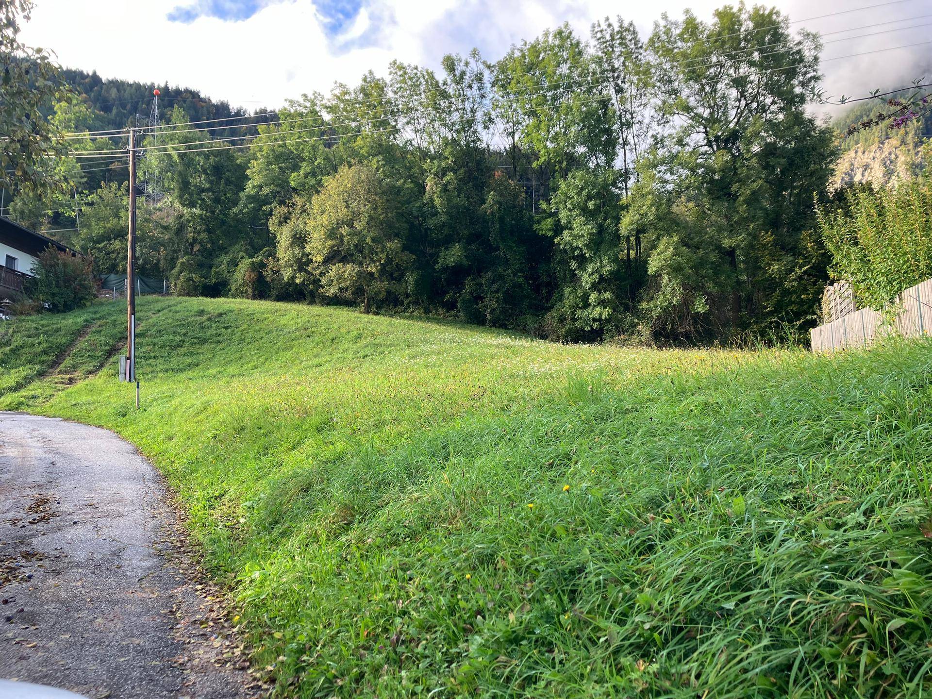Grünes, hügeliges Grundstück mit einem asphaltierten Weg und Bäumen im Hintergrund unter bewölktem Himmel.
