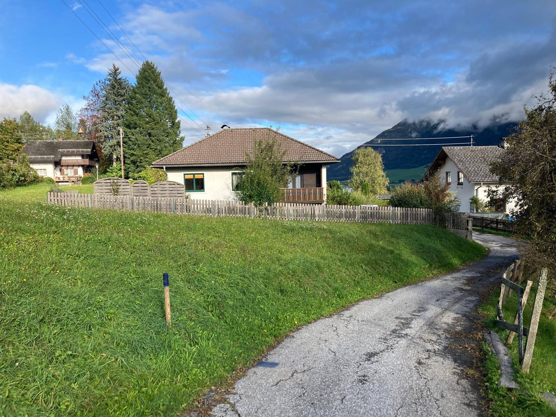 Einfamilienhaus mit braunem Ziegeldach und kleinem Balkon auf einem grünen Hügel mit Bergkulisse.