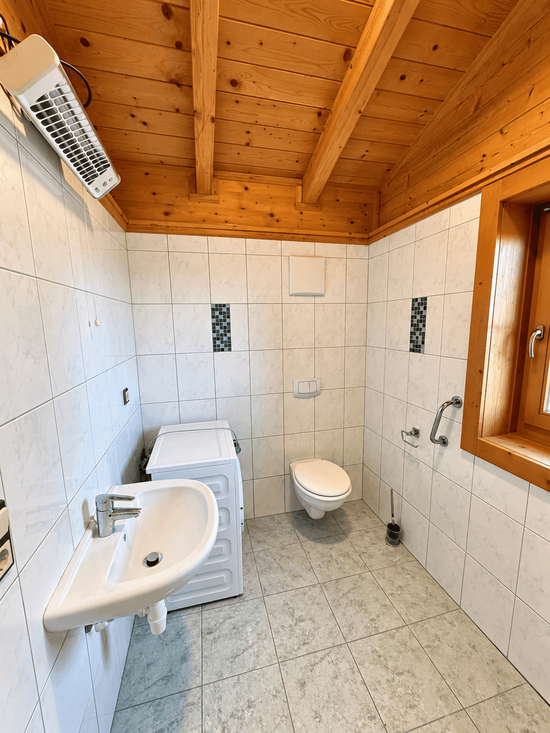 Helles Badezimmer mit weißen Fliesen, Waschbecken, Toilette und Waschmaschine.