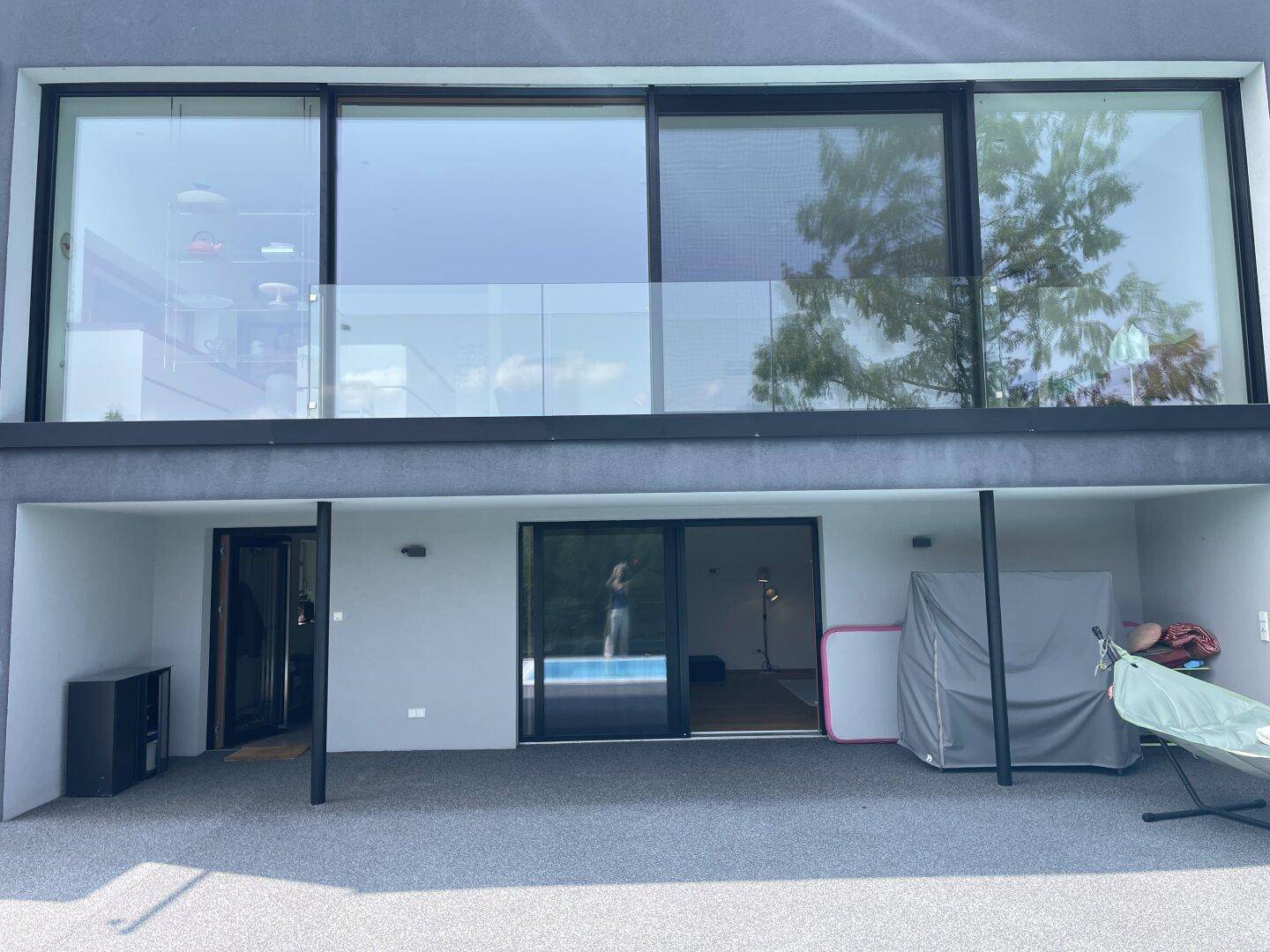 Moderne Hausfassade mit großen Fenstern und überdachtem Außenbereich, ideal für Schatten an sonnigen Tagen.