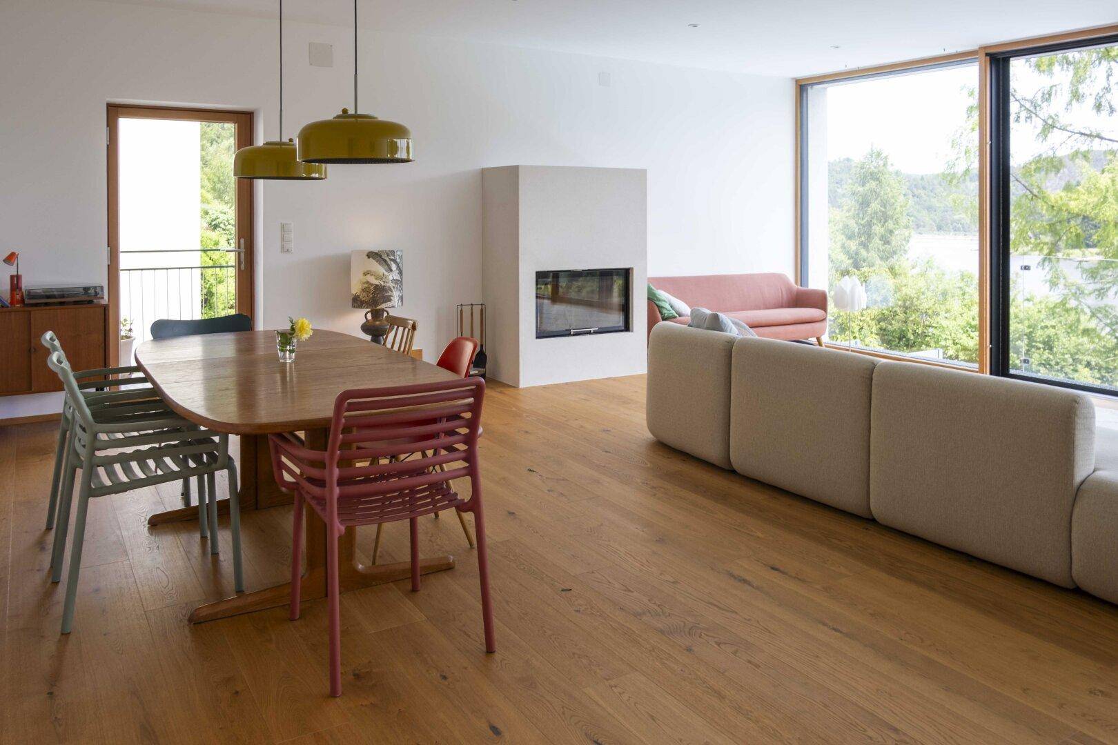 Heller offener Wohn-Essbereich mit Holzboden, Kamin und großen Fenstern mit Blick ins Grüne.