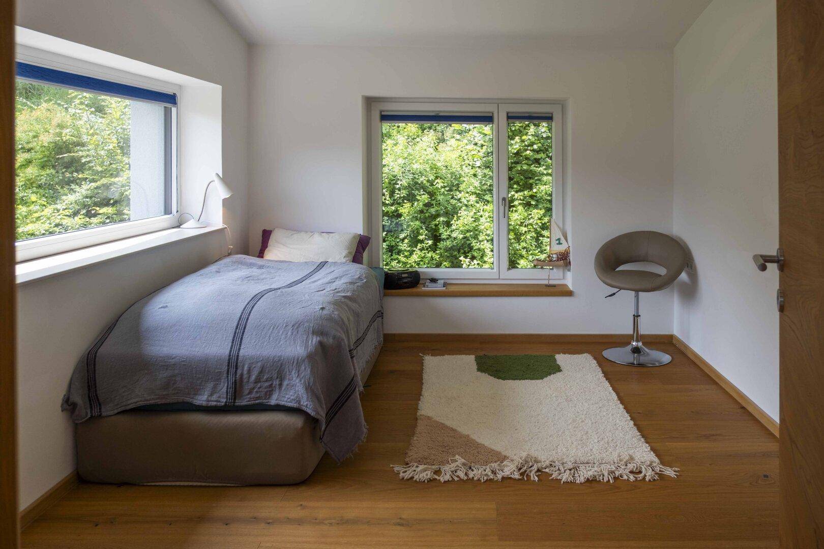 Helles Schlafzimmer mit Einzelbett, Holzboden und großen Fenstern mit Blick auf die grüne Umgebung.