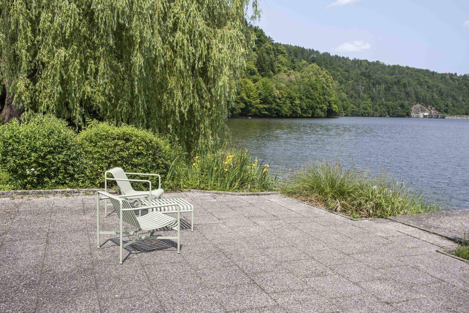 Kleine gepflasterte Terrasse im Garten mit modernen Stühlen und Blick auf den See, umgeben von Bäumen.