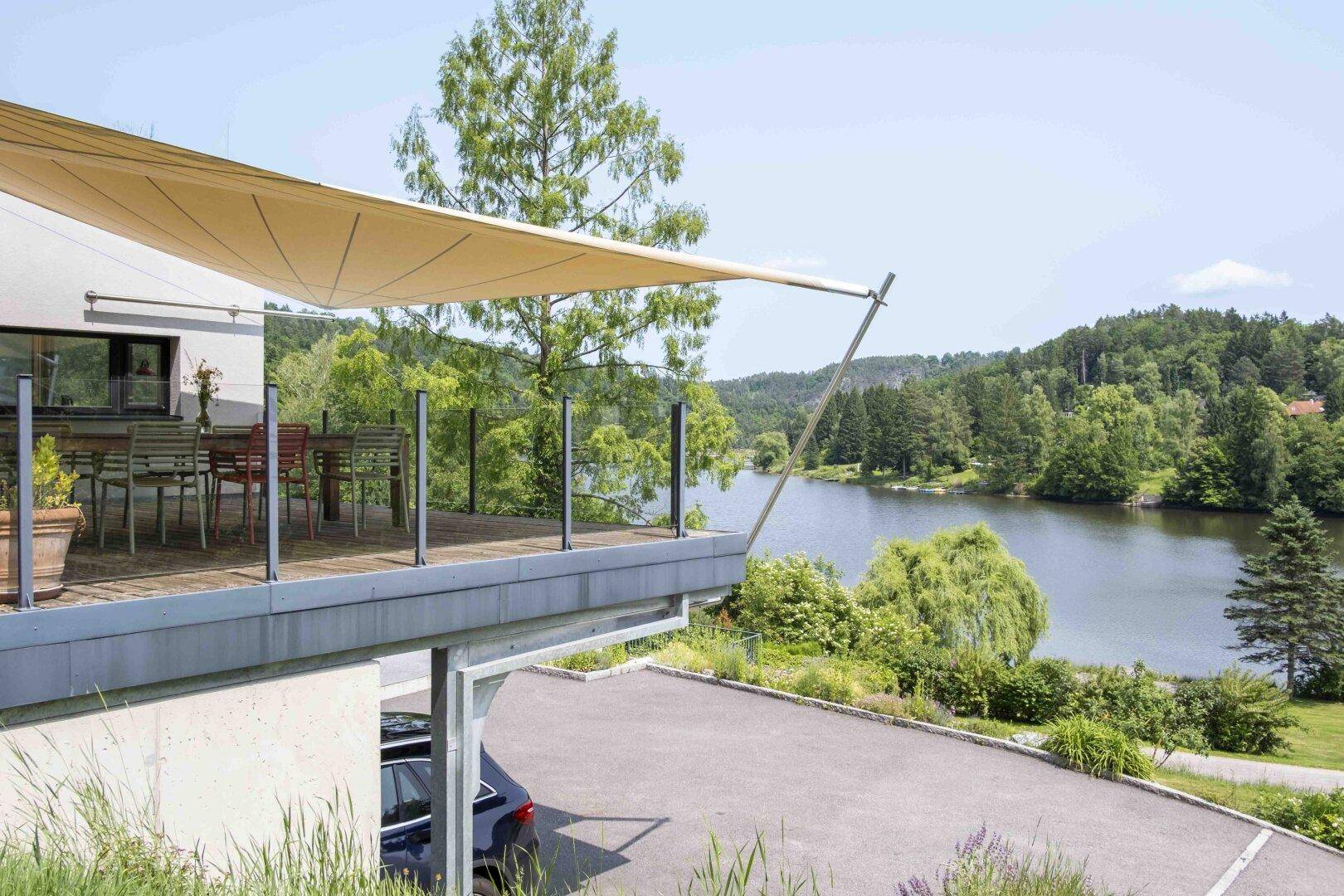 Moderne Terrasse mit Sonnensegel und Glasgeländer, bietet einen weiten Blick auf den See und die umliegende Natur.