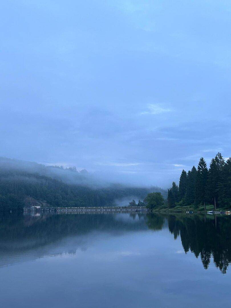 Nebliger Morgen über dem See mit bewaldeten Hügeln und einer Brücke im Hintergrund.