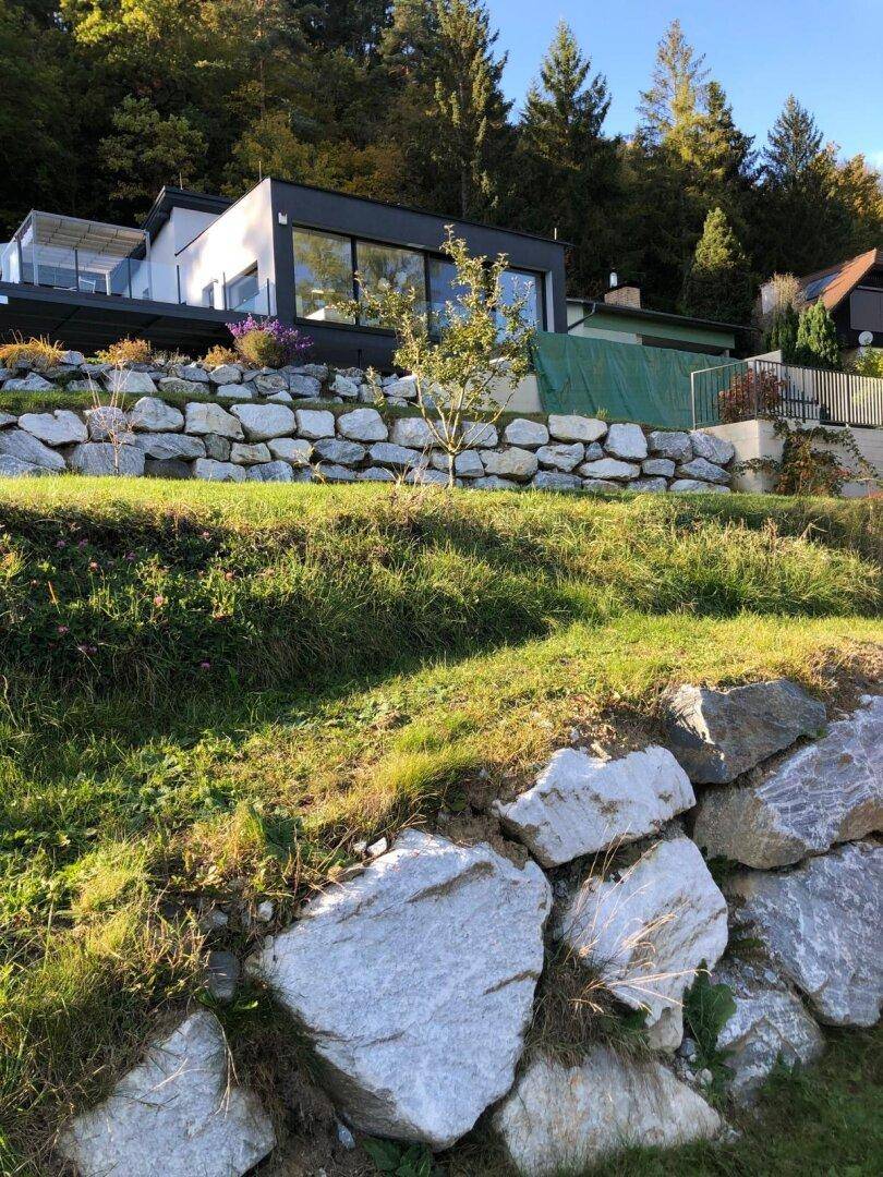 Modernes Haus am Hang mit Steingarten und dichtem Wald im Hintergrund unter blauem Himmel.