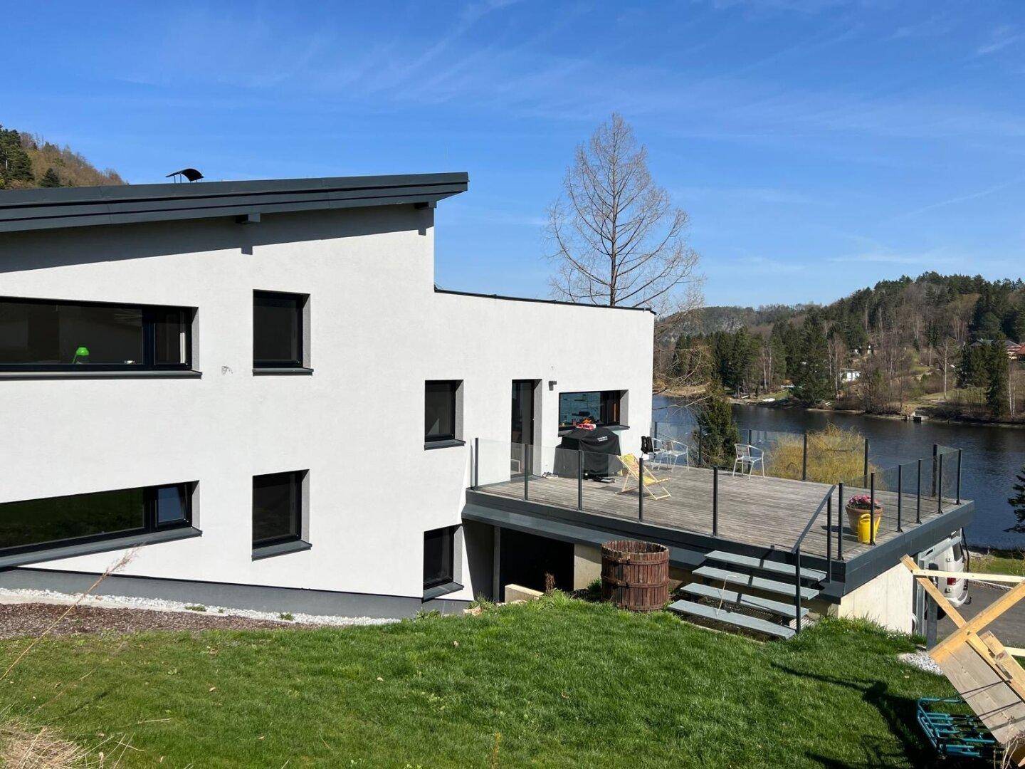 Weißes modernes Haus mit großer Holzterrasse und Grillbereich, bietet einen weiten Blick auf den See.