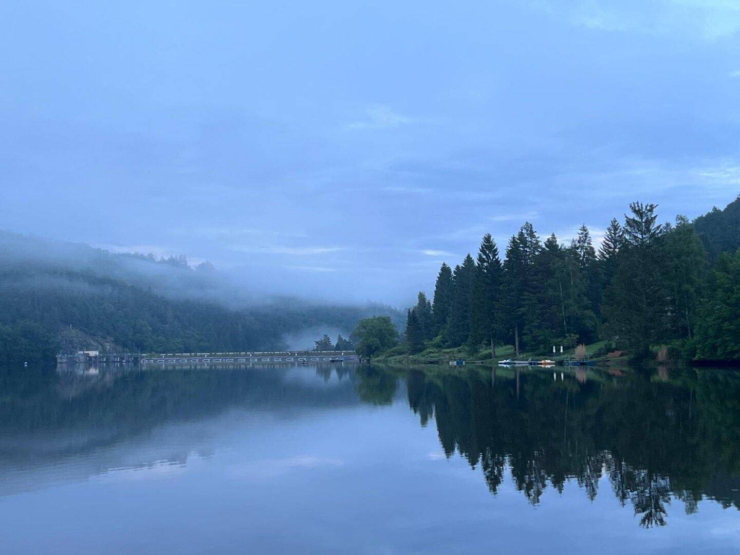 Nebliger Morgen über dem See mit bewaldeten Hügeln und einer Brücke im Hintergrund.