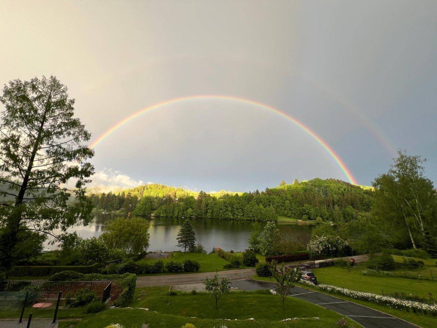 Regenbogen über dem See mit grünen Wäldern und Wiesen, eine farbenfrohe Naturerscheinung.
