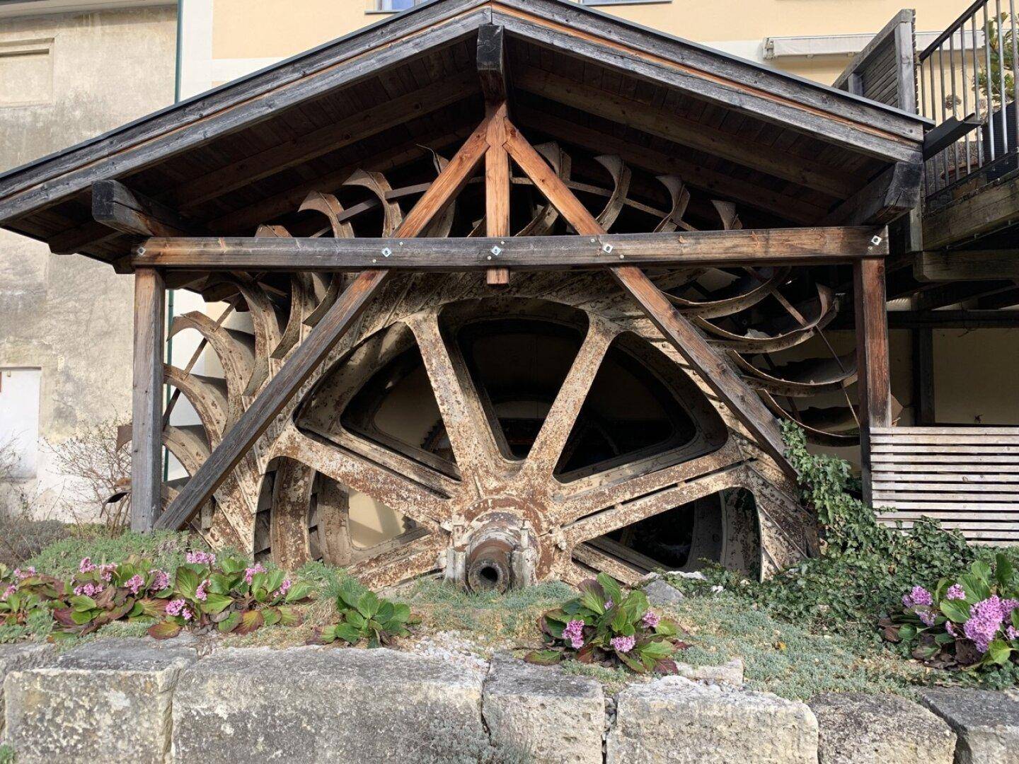 Historisches Mühlrad aus Metall und Holz, umgeben von Pflanzen.