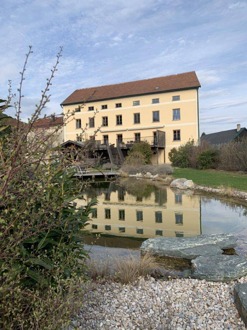 Historisches Gebäude mit gelber Fassade, umgeben von einem Teich und grüner Landschaft.