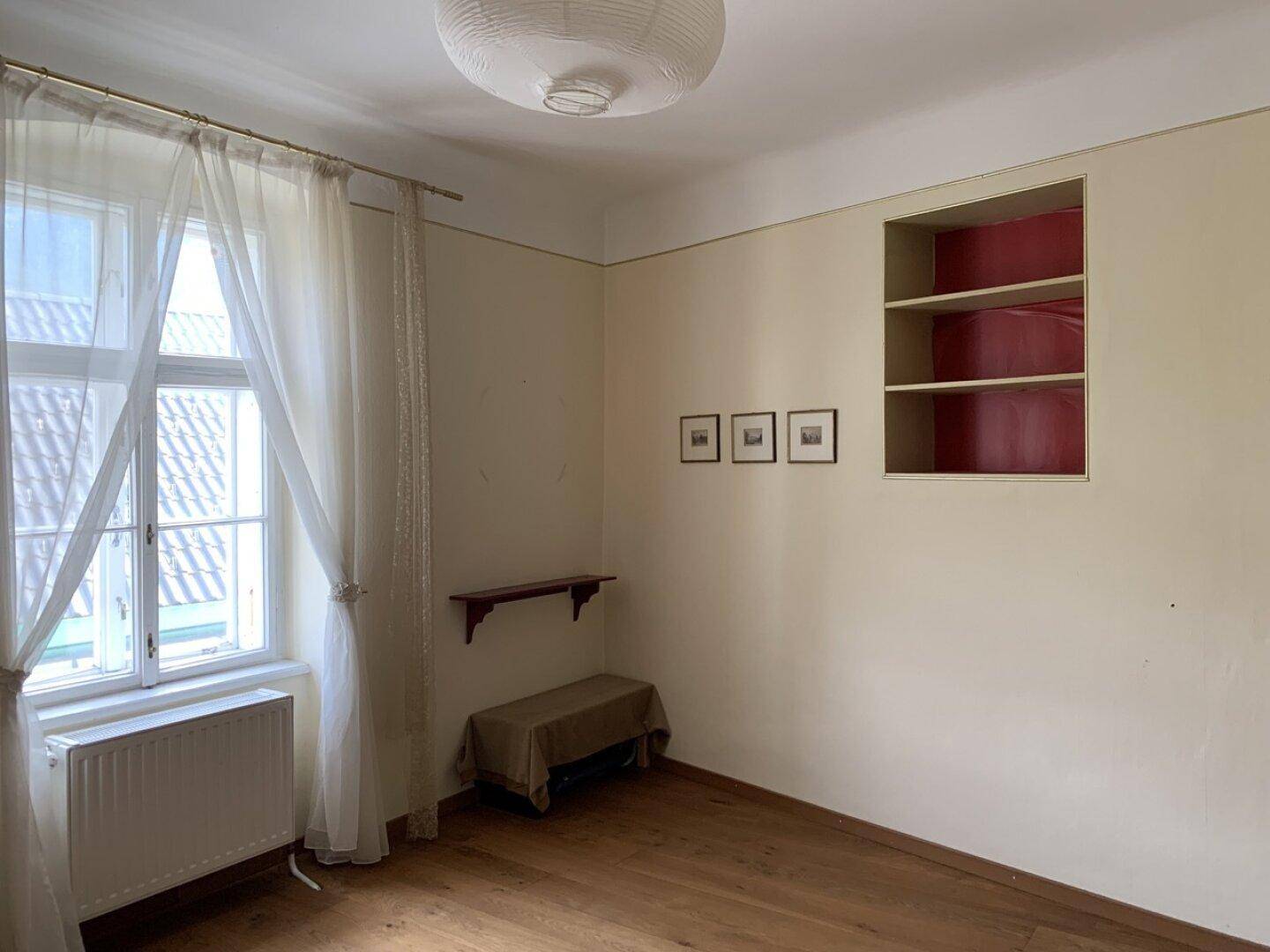 Einfaches Schlafzimmer mit Holzboden, Fenster und einem Heizkörper.