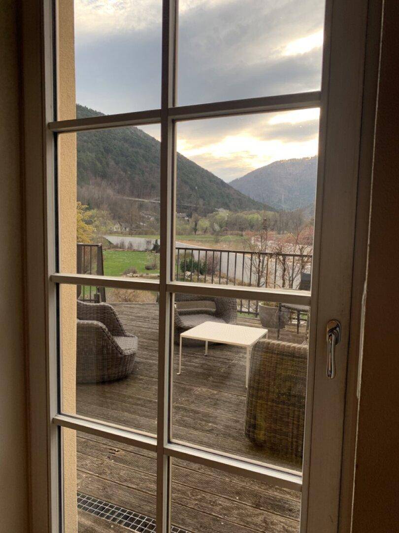 Blick aus dem Fenster auf eine möblierte Terrasse und die umliegende Berglandschaft.