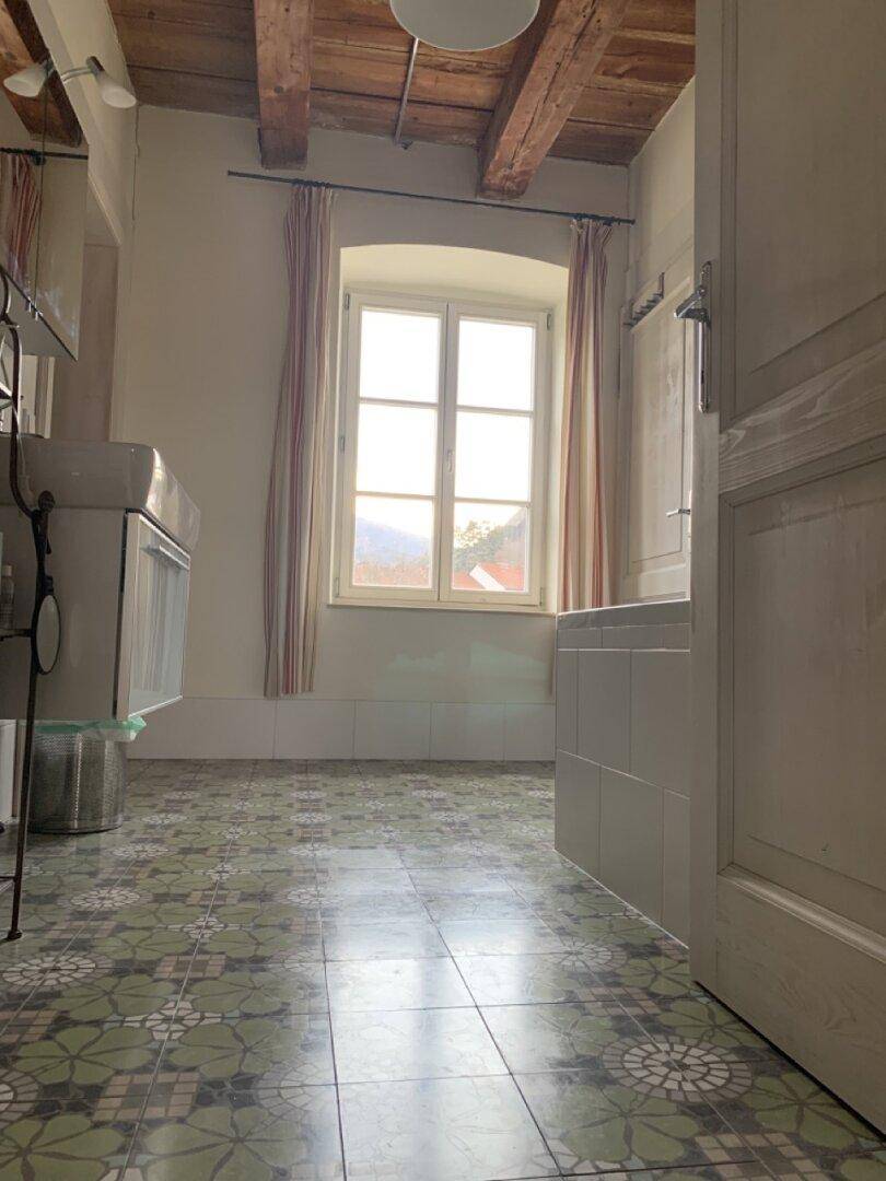 Geräumiges Badezimmer mit gemusterten Bodenfliesen, Fenster und Holzbalkendecke.
