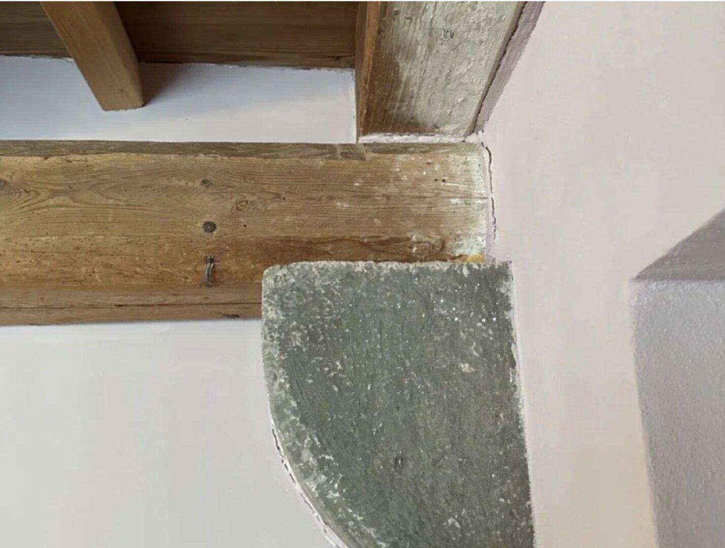 Detailaufnahme von Holzbalken und einem steinernen Stützelement in einem Innenraum.