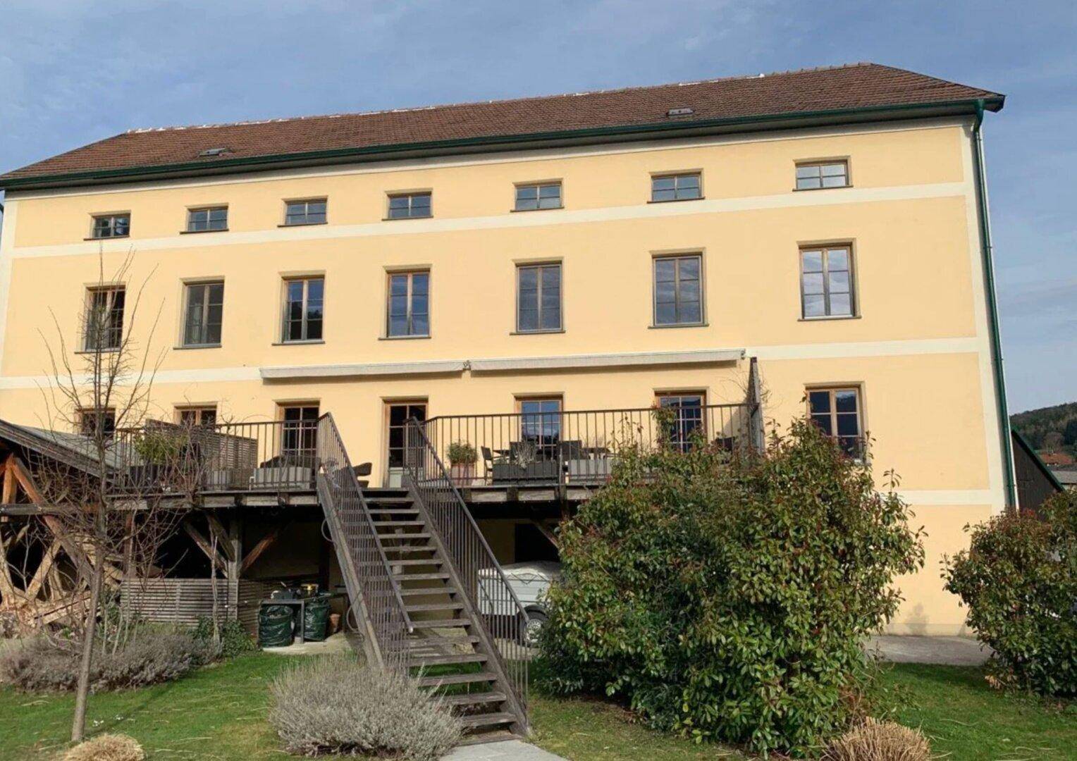 Gelbes Gebäude mit großer Holzterrasse, Außentreppe und gepflegtem Gartenbereich.