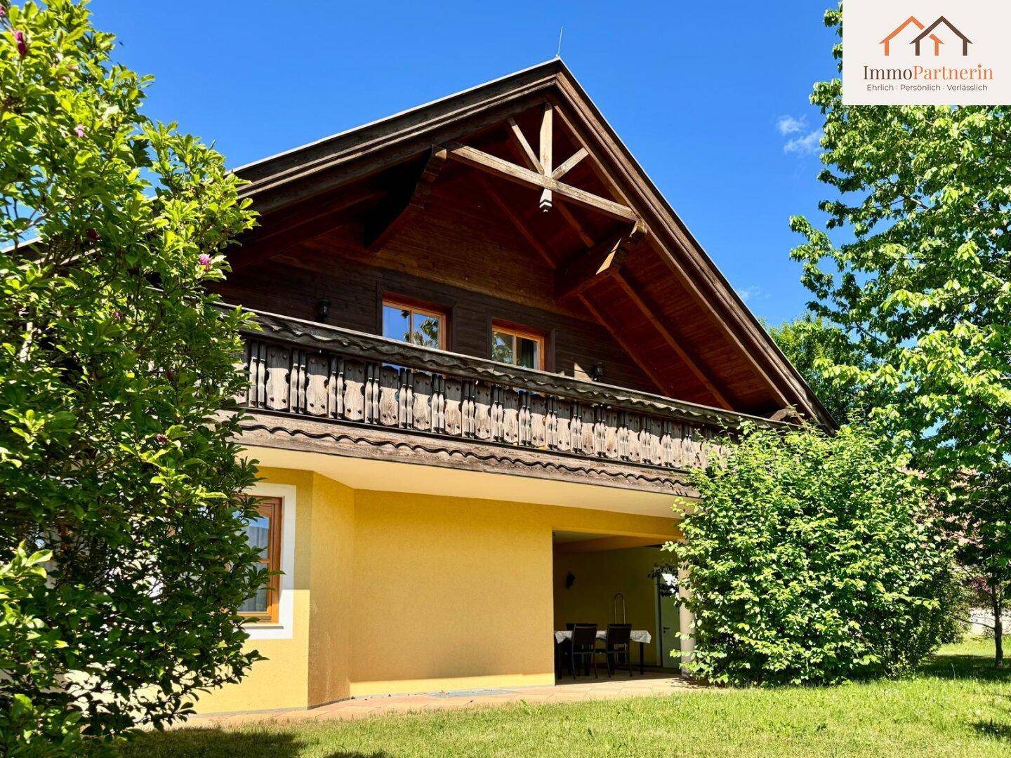 Traditionelle Hausfassade mit Holzbalkon und überdachtem Terrassenbereich, umgeben von grünen Büschen.