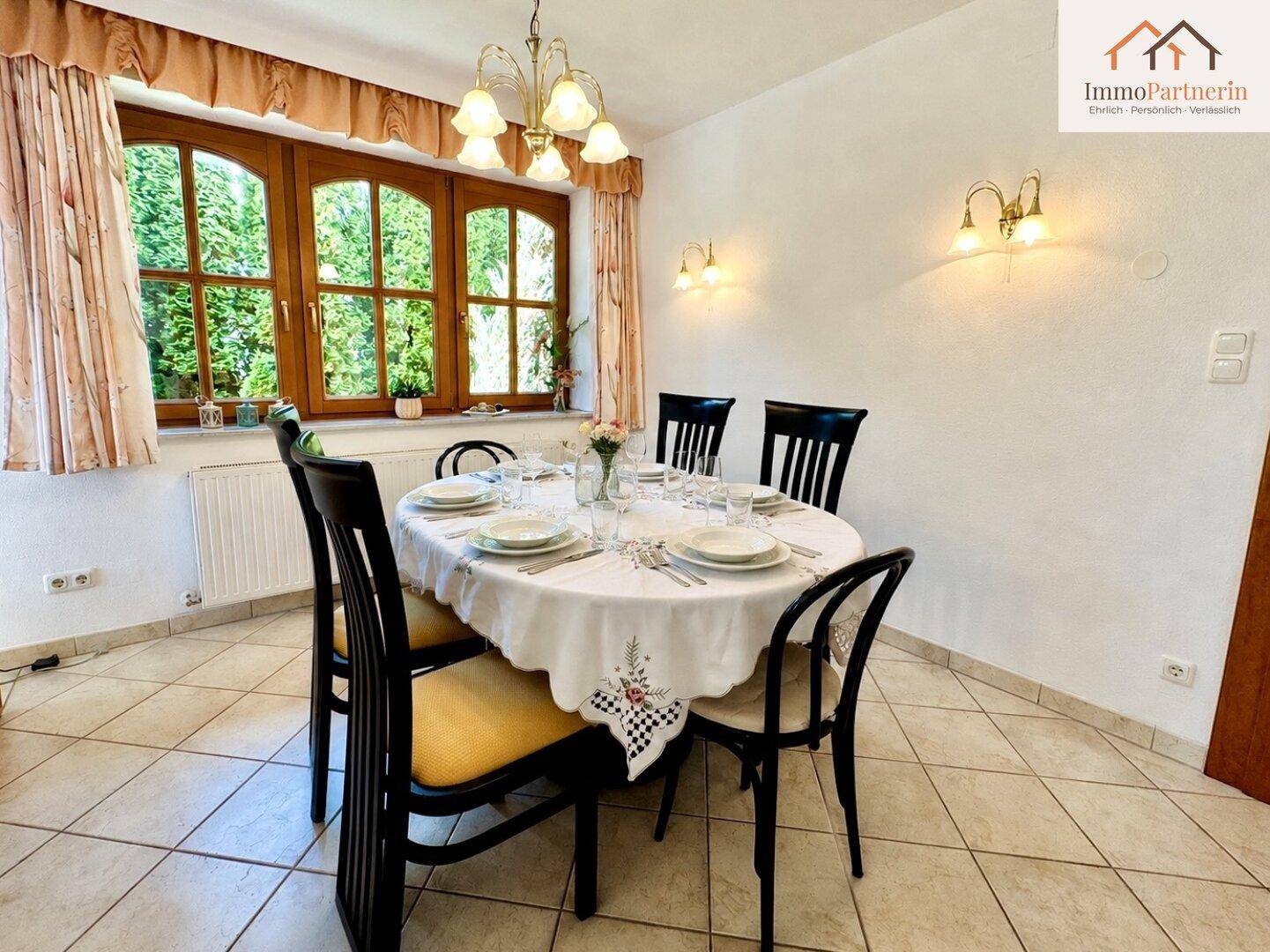 Heller Essbereich mit großem ovalen Tisch, Fliesenboden und großen Fenstern mit Blick ins Grüne.