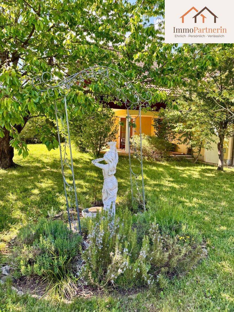 Gepflegter Gartenbereich mit einer dekorativen Statue unter einem Metallbogen und üppiger Bepflanzung.